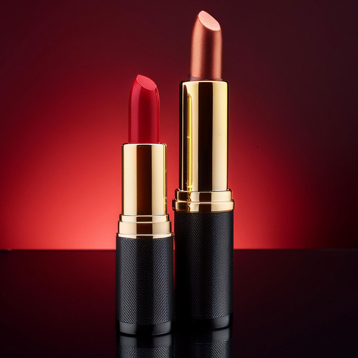 Packshot luxe cosmétique : deux rouges à lèvres dorés et noirs sur fond rouge dégradé, reflets maîtrisés